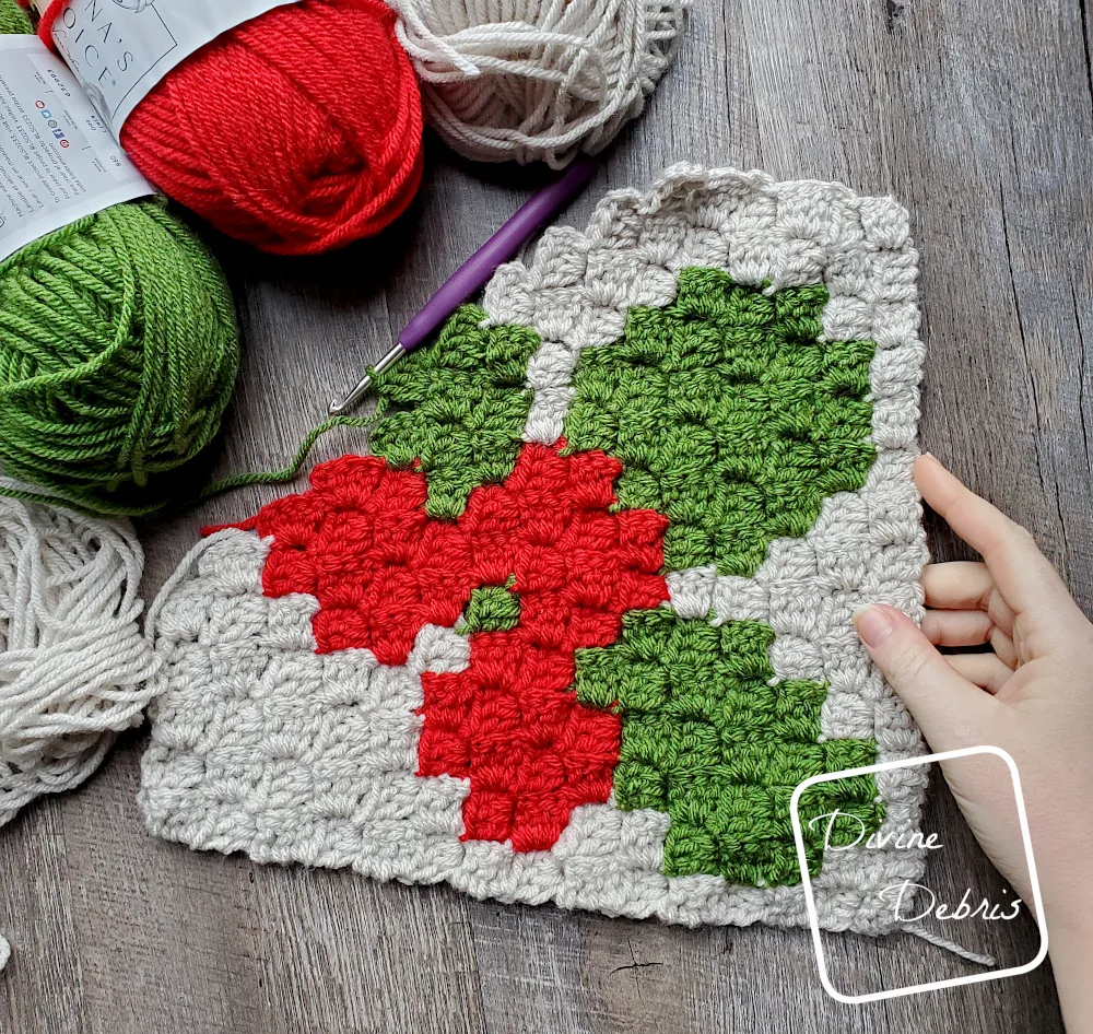 [Image description] The unfinished C2C Winter Holly Afghan Square laying on a wood grain background with skeins of yarn on the top left and a white woman's hand holding the bottom right corner.