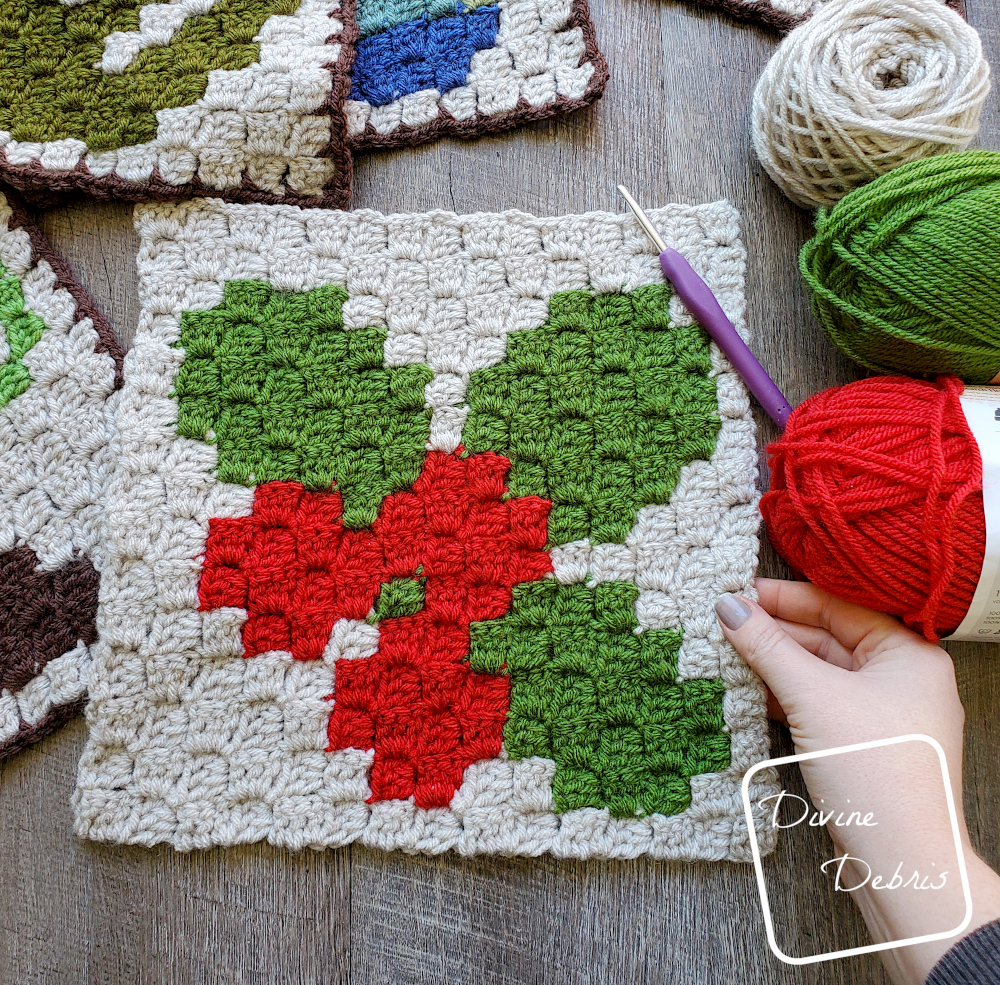 [Image description] The C2C Winter Holly Afghan Square laying on a wood grain background with skeins of yarn to the right and a white woman's hand holding the bottom right corner.