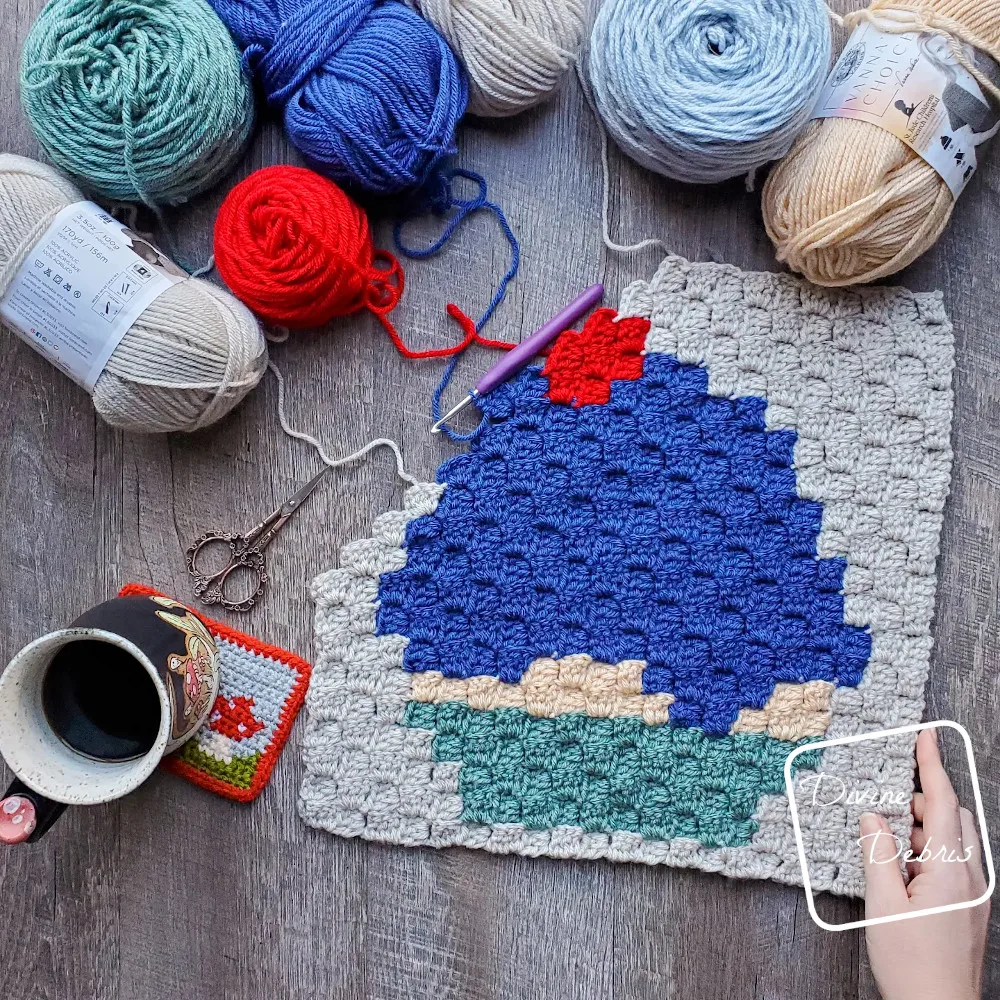 [Image description] A top down look at an unfinished C2C Cupcake Afghan Square with skeins of yarn on the left top side and a white woman's hand holding the right bottom corner