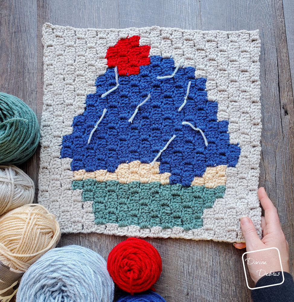 [Image description] A top down look at the C2C Cupcake Afghan Square with skeins of yarn on the left bottom side and a white woman's hand holding the right bottom corner