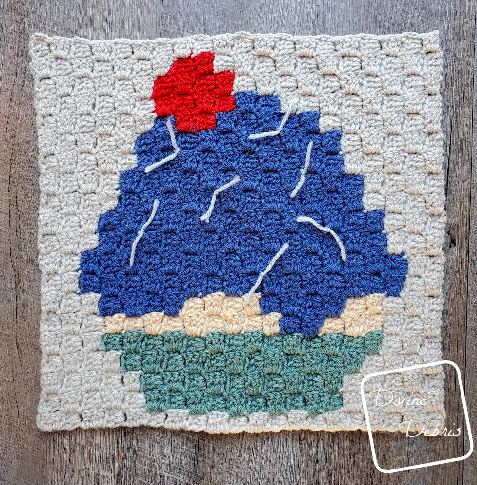 [Image description] A top down flat lay of the C2C Cupcake Afghan Square with blue frosting, a red cherry on top and light blue sprinkles