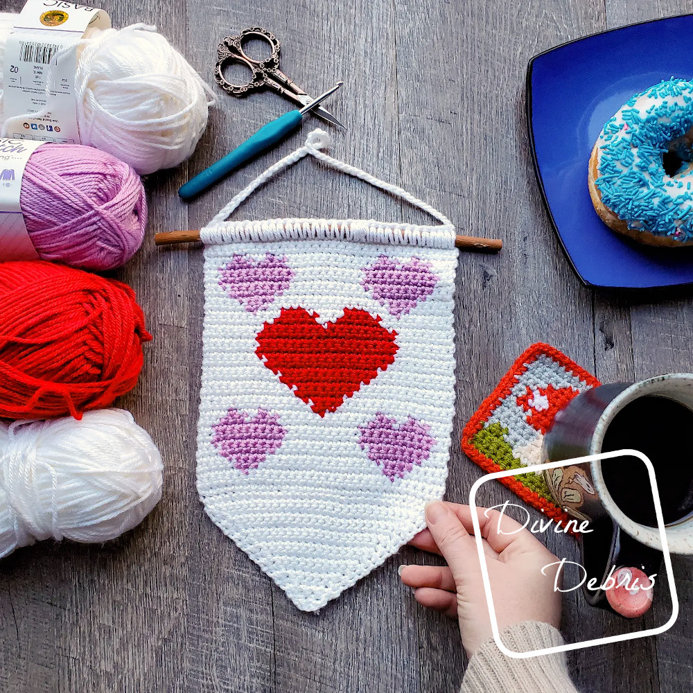 [Image description] The Cute Hearts Wall Hanging lays flat on a fake wooden background, a white woman's hand holds the bottom right side, skeins of yarn are on the left and a donut and cup of coffee are on the right.