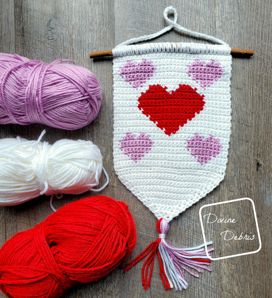 [Image description] Cute Hearts Wall Hanging lays on a fake wood background with 3 skeins of yarn on the left side.