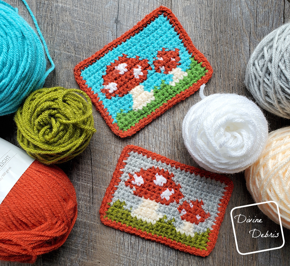 [Image description} 2 versions of the Cute Mushrooms Mug Rug lay flat on a wood grain background surrounded by skeins of yarn