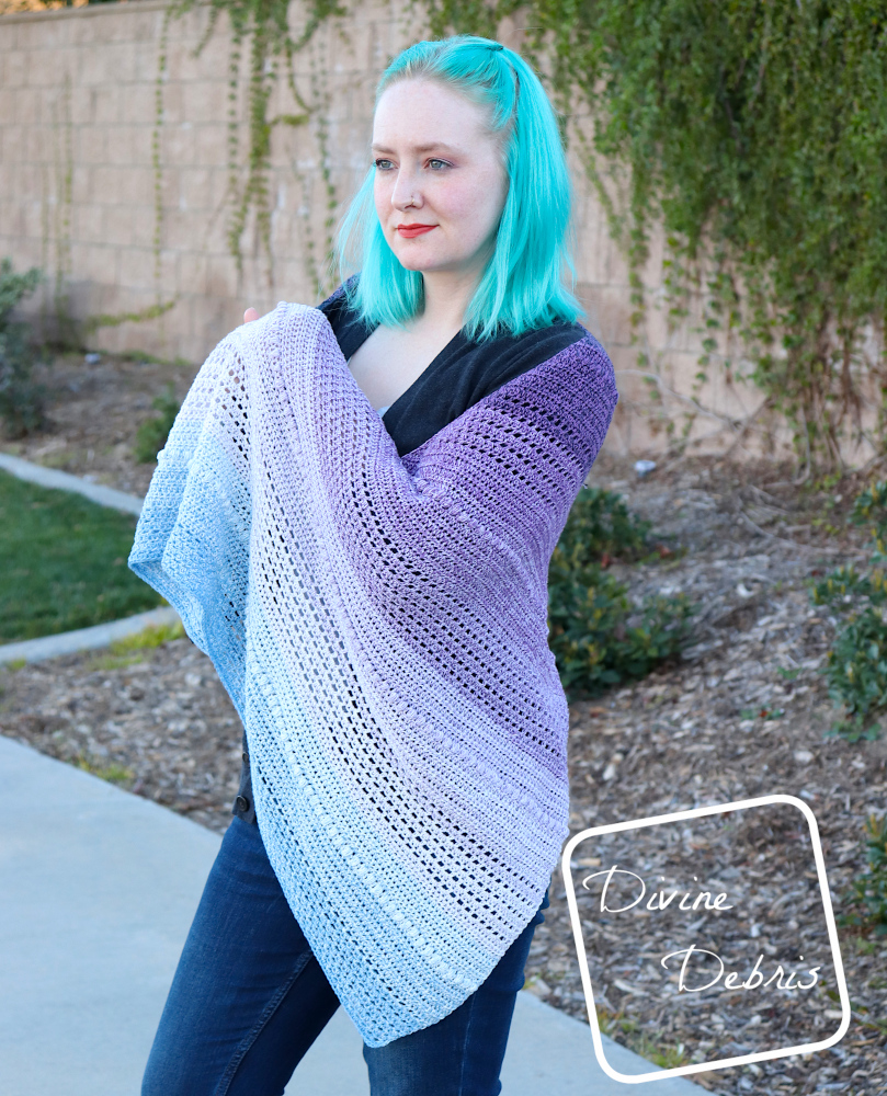 [Image description] A white woman with blue hair stands looking away from the camera holding the Ursula Shawl in front of a tan brick wall with ivy 