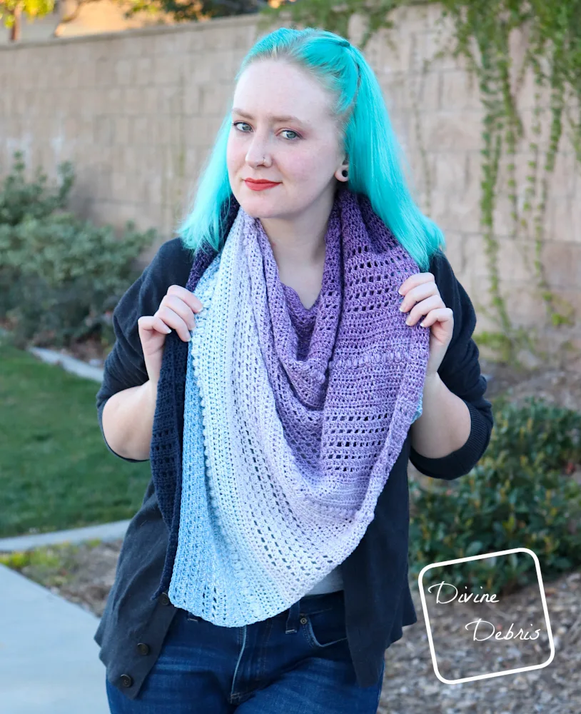 [Image description] A white woman with blue hair faces the camera with the Ursula Shawl wrapped around her neck like a scarf, in front of a tan brick wall with ivy 