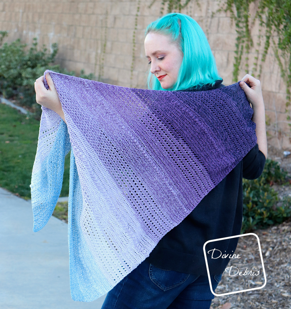 [Image description] A white woman with blue hair faces away from the camera showing off the Ursula Shawl across her back, in front of a tan brick wall with ivy 