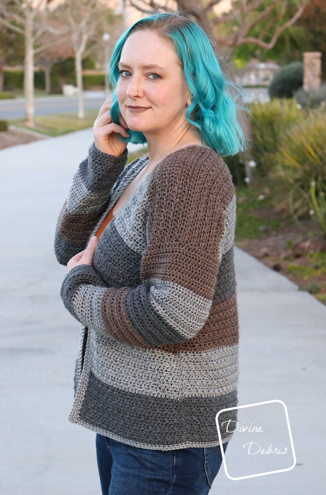[image description] Side view of a white woman with blue hair wearing the Alix Cardigan, looking at the camera standing on a road with lots of green landscaping