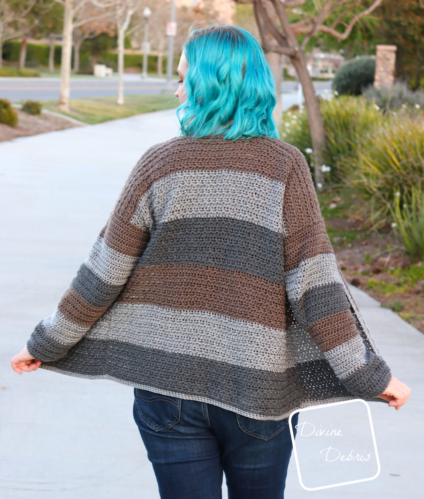 [Image description] A back view of the Alix Cardigan being worn by a white woman with blue hair on a street with a lots of greenery.