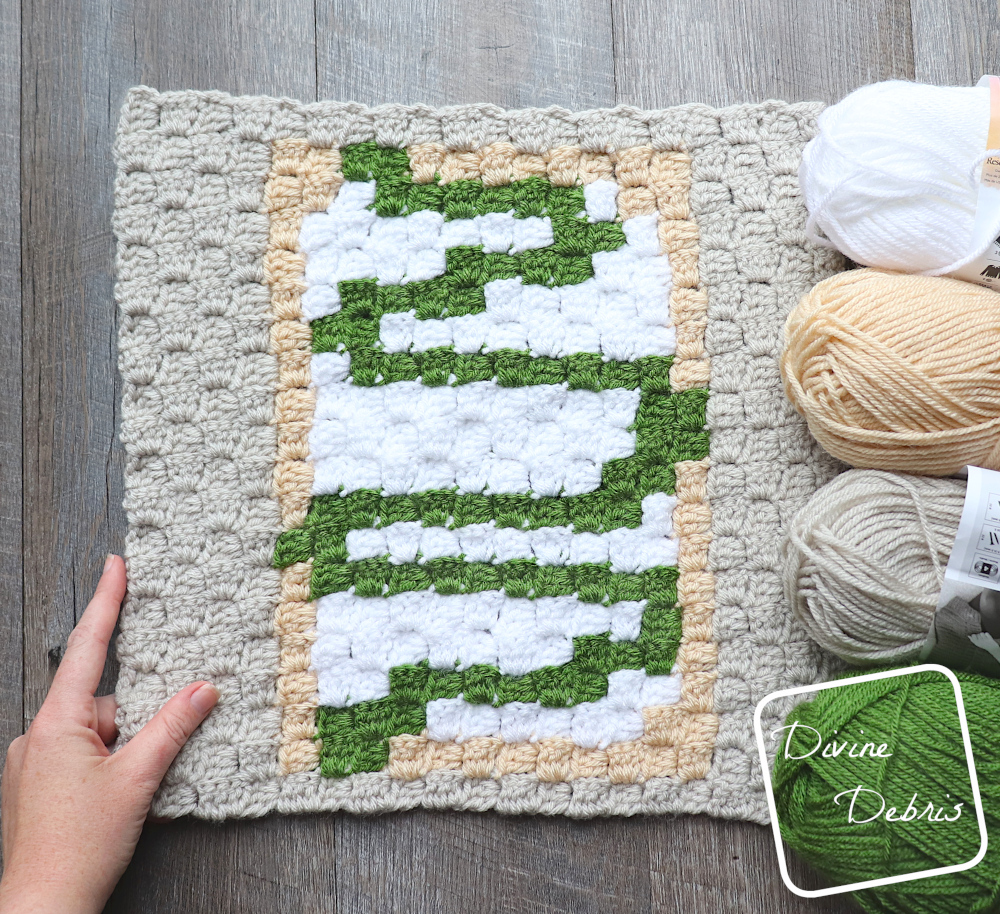 [Image description] A top down photo of the C2C Toaster Pastry Afghan Square on a fake wood background, with 4 skeins of yarn along the right side of the photo and a white woman's hand holding the bottom left corner.