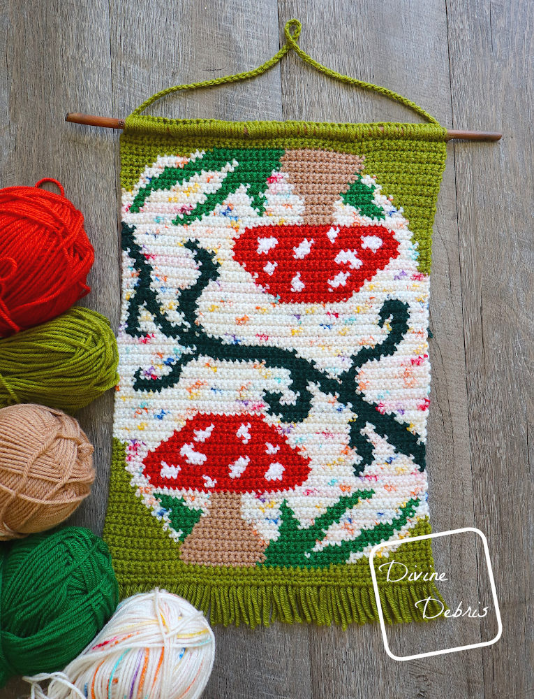 [Image descritption] Top down view of the Mushroom Reflections Wall Hanging with skeins of yarn around the bottom left of the hanging.