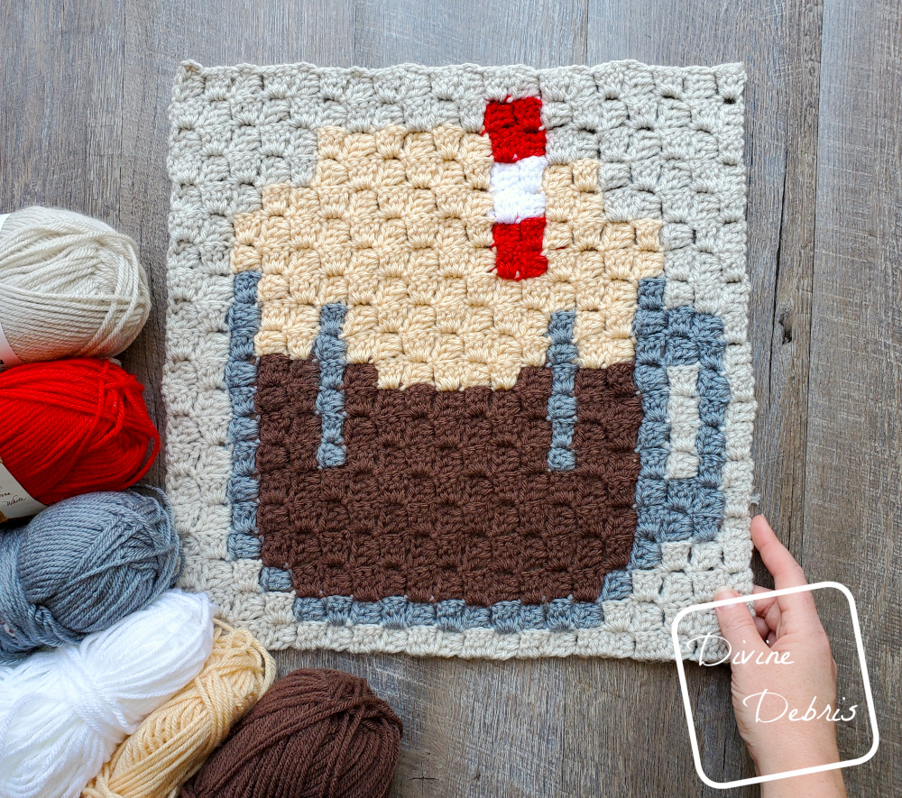 [Image description] Top down view of the the C2C Root Beer Float Afghan Square with skeins of yarn on the bottom left corner and a white woman's hand holding the bottom right corner.