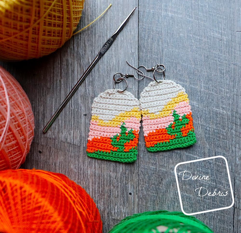 [Image description] Top down view of the Sedona Cactus Crochet Earrings laying on a wood grain background with skeins of crochet thread bordering it to the bottom and left.