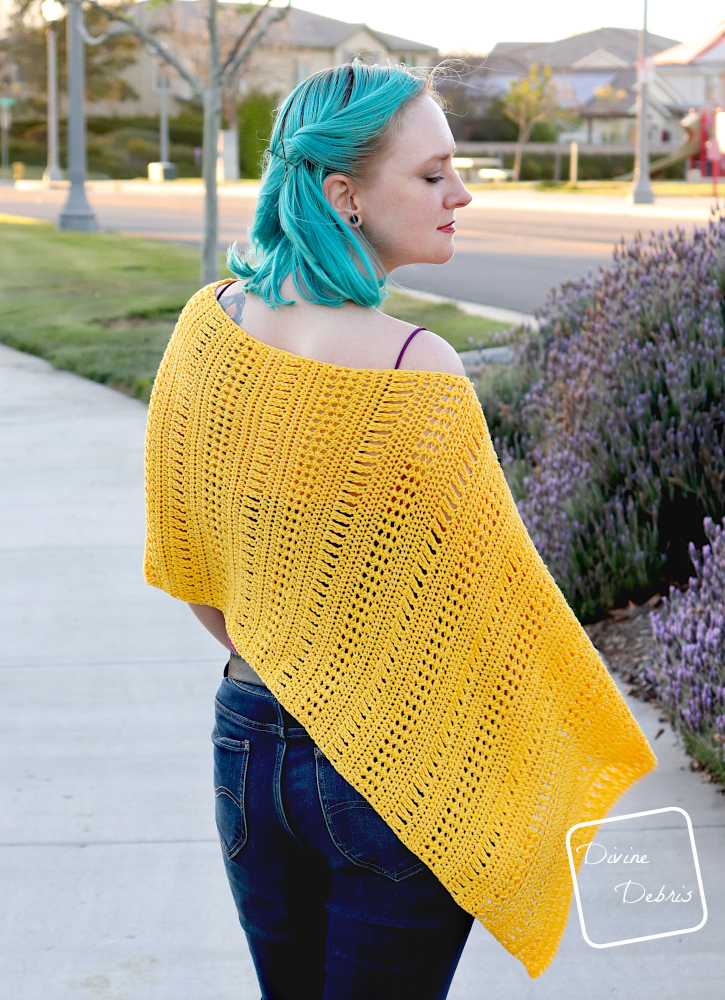 [Image description] A white woman with blue hair stands facing away from the camera on a sidewalk wearing the yellow Stephanie Poncho