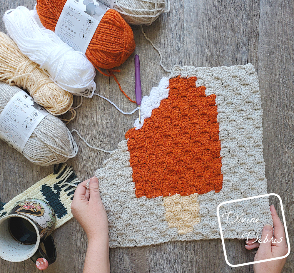 [Image description] Top down view of an unfinished Orange Creamsicle Afghan Square, a white woman's hand holds the bottom corners and 5 skeins of yarn sit along the top left of the photo