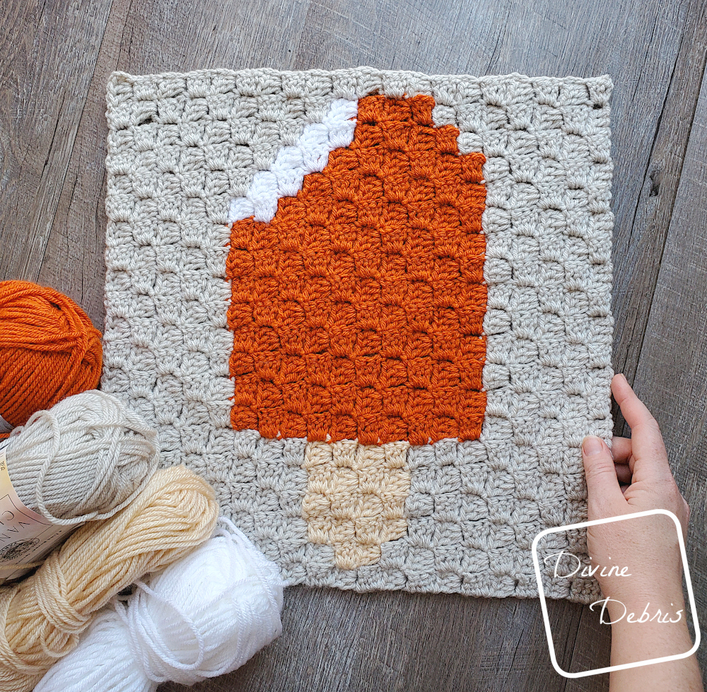 [Image description] Top down view of the Orange Creamsicle Afghan Square sitting on a wood grain background, a white woman's hand holds the bottom right corner and 4 skeins of yarn sit on the left bottom corner.