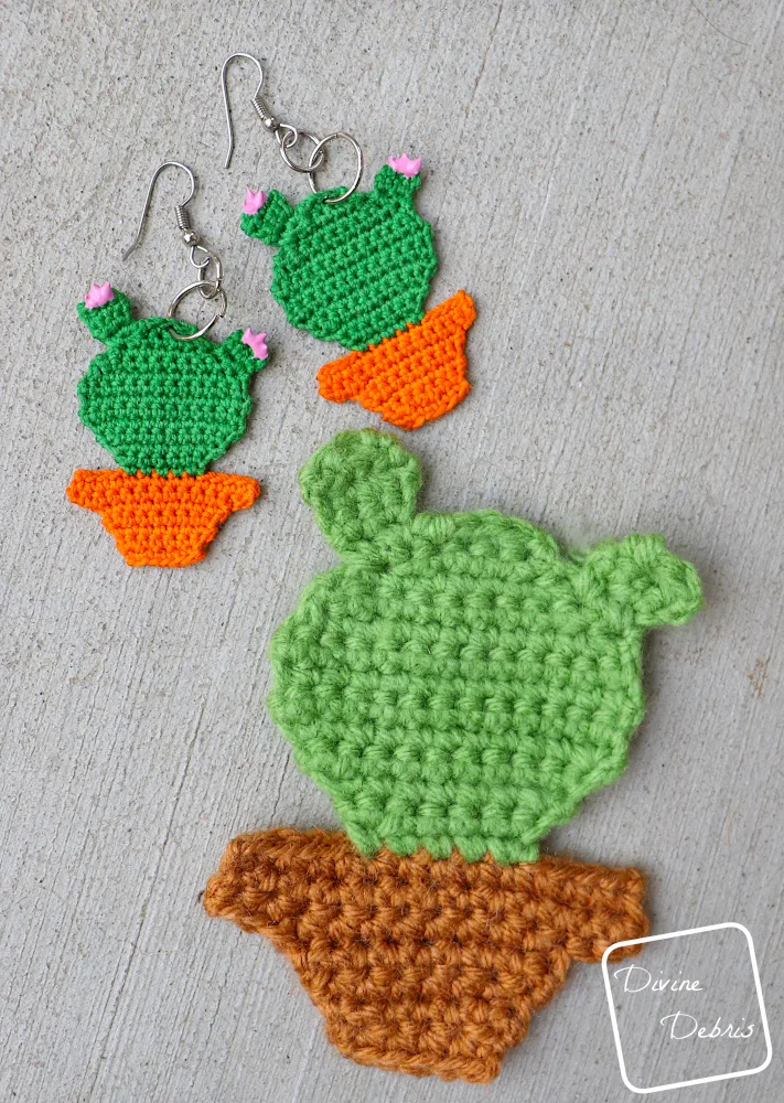 [Image description] A pair of the Cute Cactus Earrings and an applique Cute Cactus lay next to each other on a cement background.