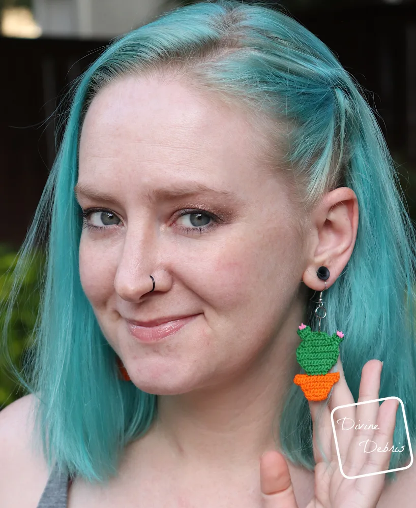[Image description] A white woman white blue hair looks directly at the camera white holding the Cute Cactus Earring to face the camera
