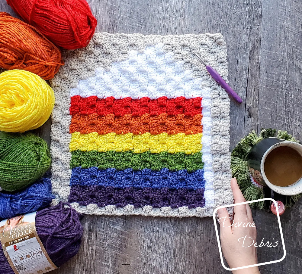 [Image description] C2C Rainbow Cake Afghan Square laying flat on a wood grain background with skeins of yarn on the left side and a white woman's hand holding the bottom right corner
