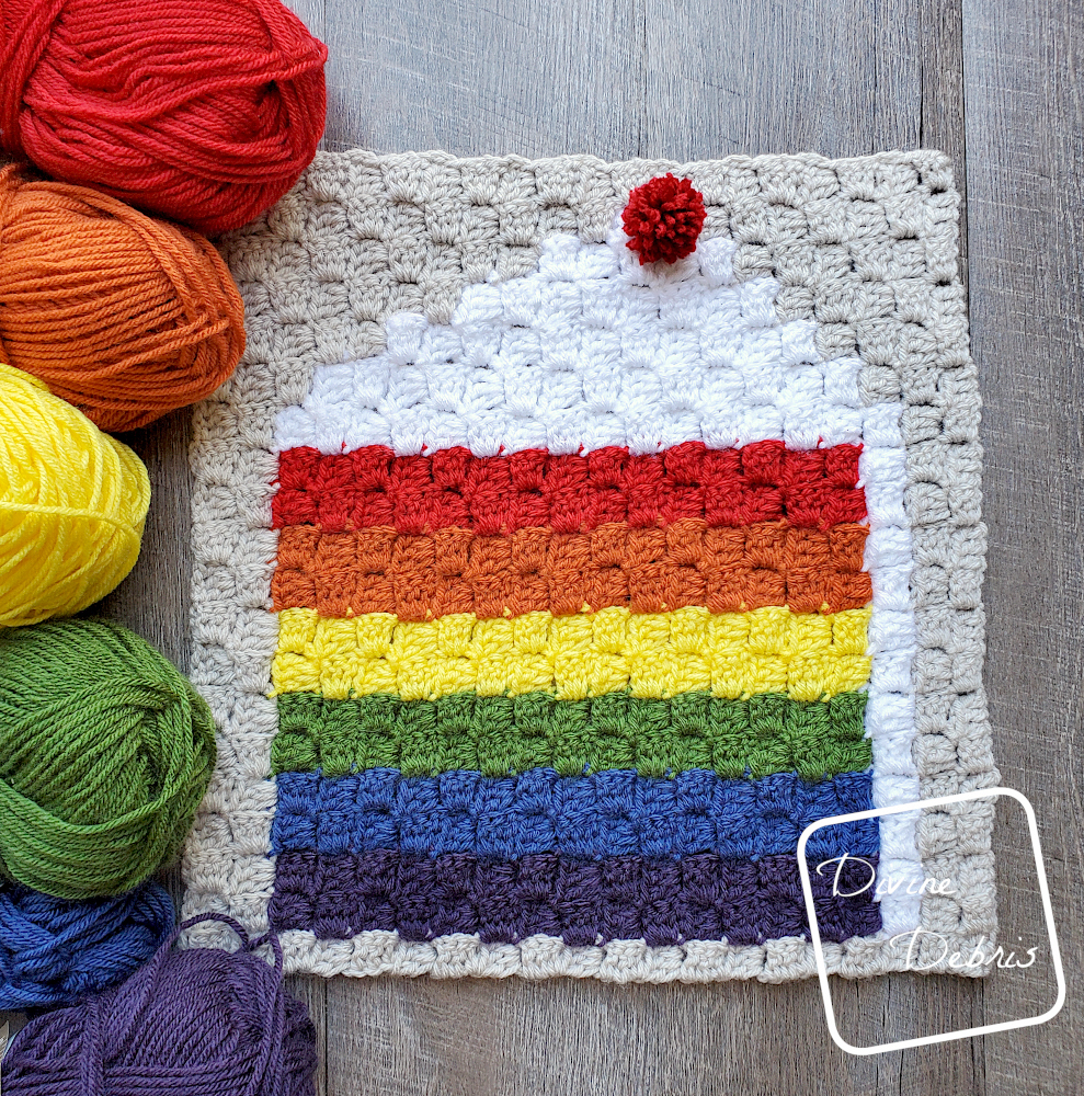 [Image description] C2C Rainbow Cake Afghan Square laying flat on a wood grain background with 6 skeins of yarn, in a rainbow arrangement, along the left side of the square