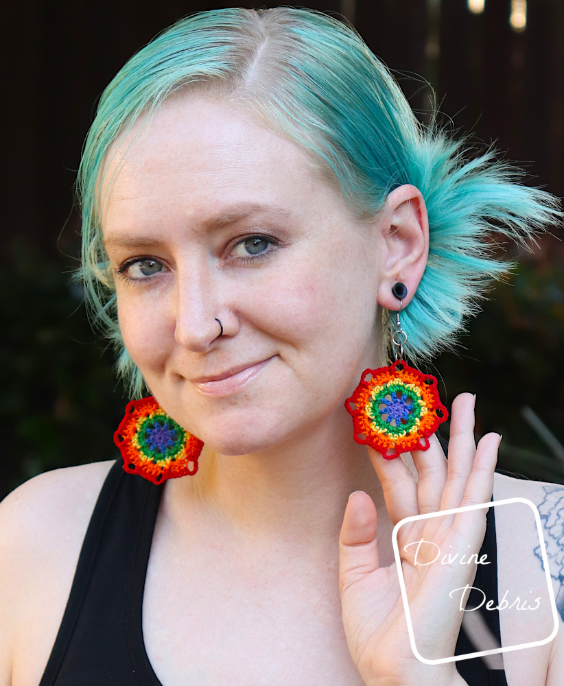 [Image description] A white woman with blue hair stands facing the camera, hand holding the back of her earring to the camera.