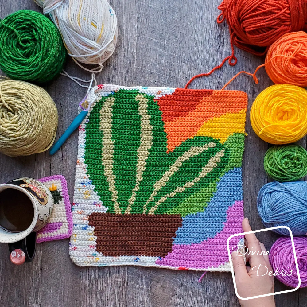 [Image description} Top down view of the unfinished Rainbow Cactus Wall Hanging on a wood grain background with skeins of yarn on both sides and a white woman's hand holding the bottom right corner.