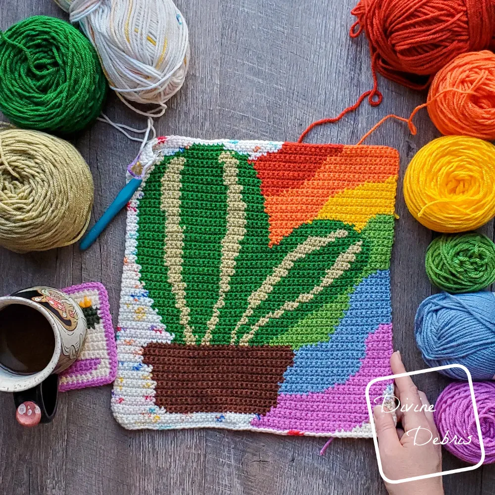 [Image description} Top down view of the unfinished Rainbow Cactus Wall Hanging on a wood grain background with skeins of yarn on both sides and a white woman's hand holding the bottom right corner.