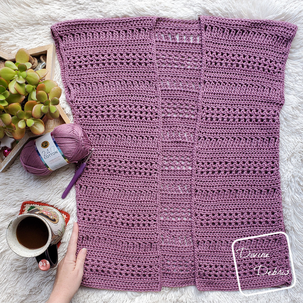 [Image description] The Stephanie Cardigan crochet pattern lays flat on a white blanket with a white woman's hand holding the bottom left corner.