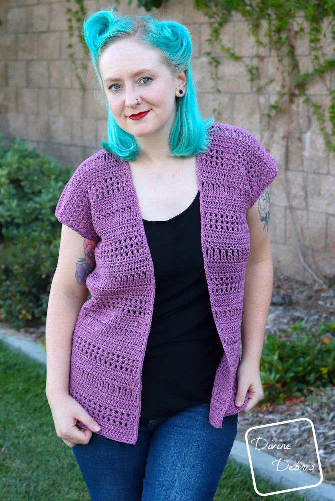 [Image description] A white woman with blue hair wearing the Stephanie Cardigan crochet pattern faces the camera with her hand at her hips, in front of a tan wall and bushes 