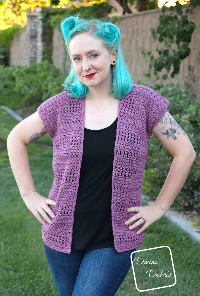[Image description] A white woman with blue hair wearing the Stephanie Cardigan crochet pattern stands with her hands on her hips facing the camera, in front of a tan wall and bushes 
