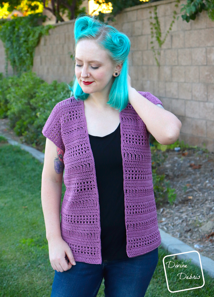 [Image description] A white woman with blue hair wearing the Stephanie Cardigan crochet pattern looks down and to the right, in front of a tan wall and bushes 