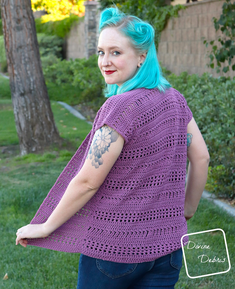 [Image description] A white woman with blue hair wearing the Stephanie Cardigan crochet pattern looking over her left shoulder at the camera, in front of a grass, a tree, and bushes