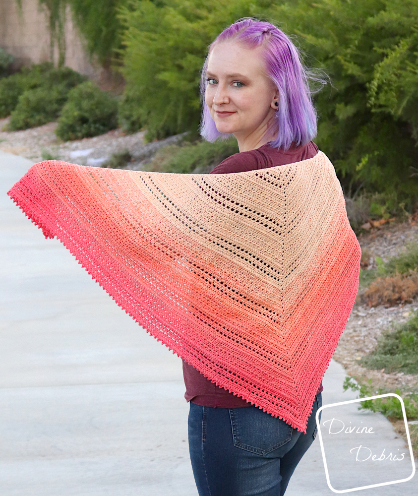 [image description] a white woman with purple hair stands on a side walk in front of bushes and a wall, facing away but holding the Alexis shawl toward the camera