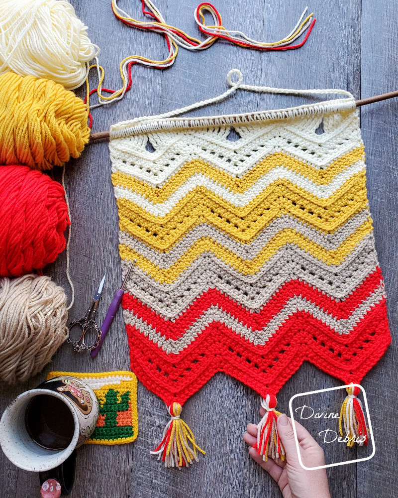 [Image description] The Colorful Chevrons Wall Hanging lays flat on a wood grain background with skeins of yarn on the left side of the frame an a white woman holds one of the tassels on the bottom. 
