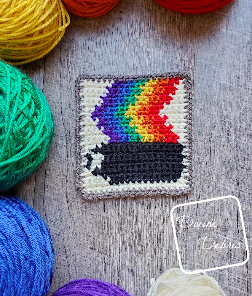 [Image description] A close up of a Rainbow Coffee Coaster sitting on a wood grain background with cakes of yarn surrounding it.