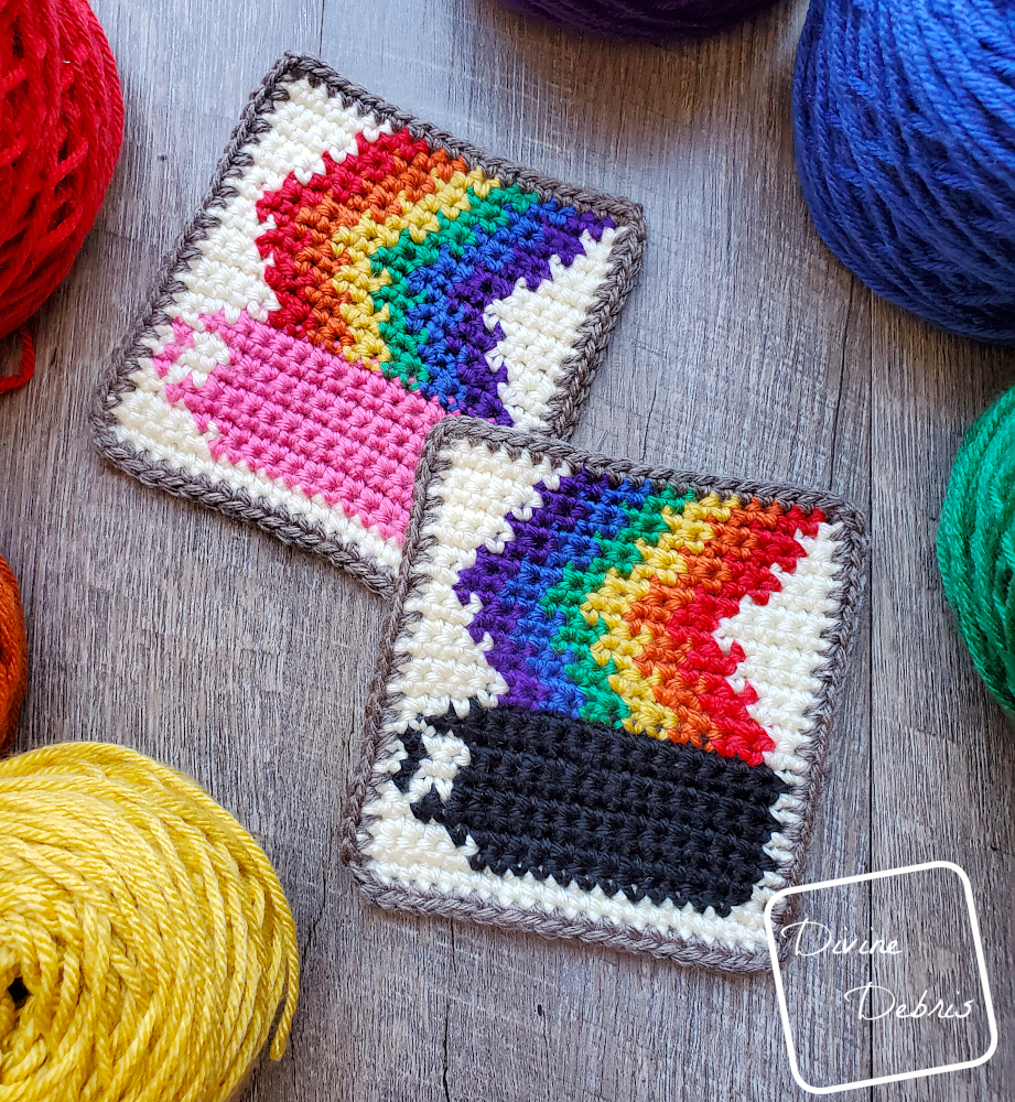 [Image description] A close up of 2 Rainbow Coffee Coasters sitting on a wood grain background with cakes of yarn surrounding them.