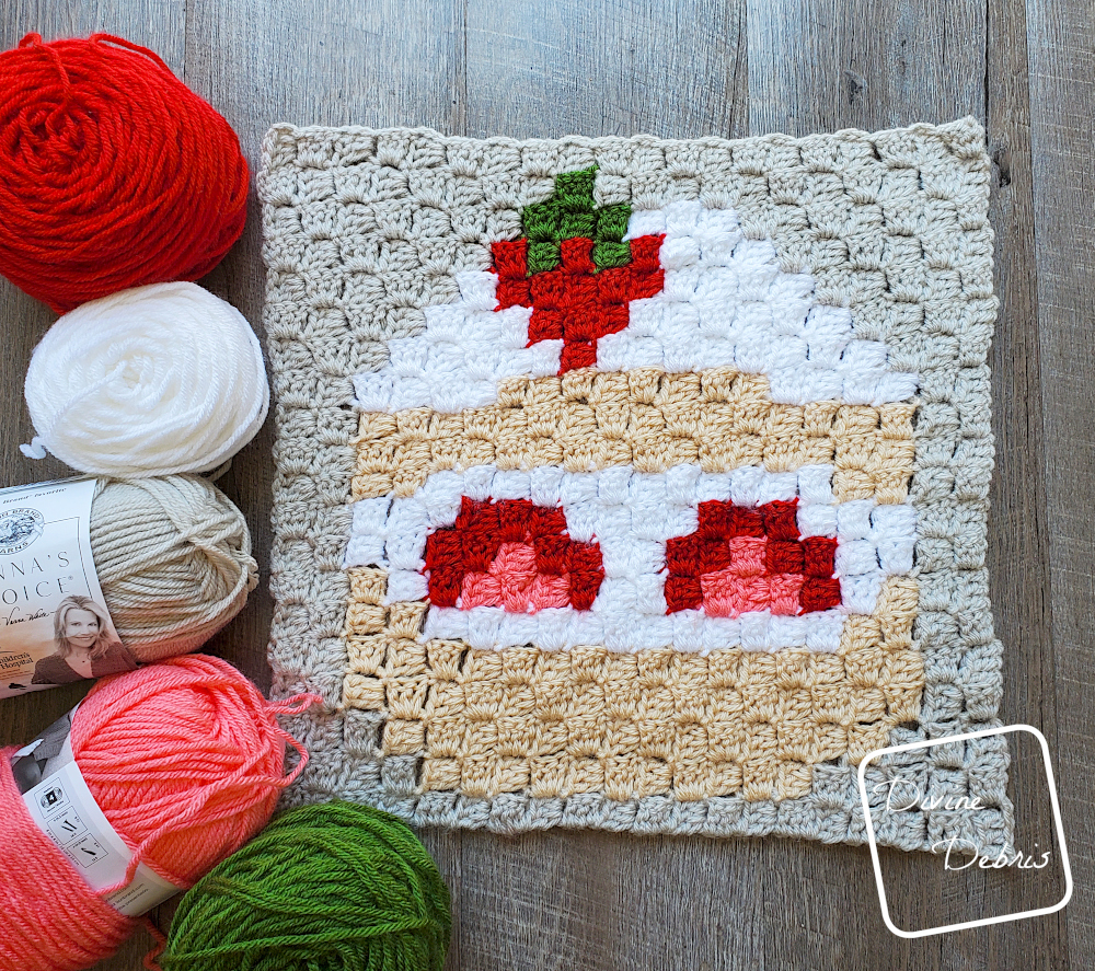 [Image description] C2C Strawberry Shortcake Afghan Square flat on a wood grain background, with skeins of yarn on the left side of the square.