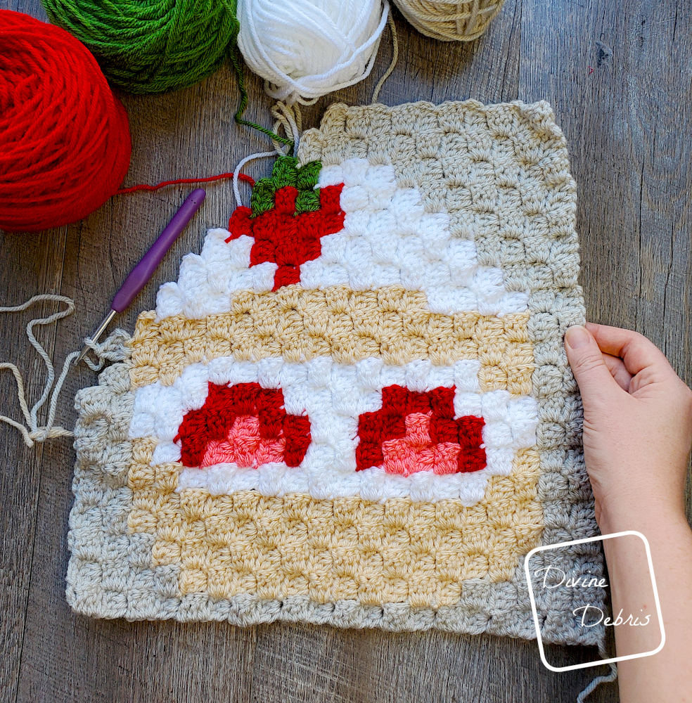 [Image description] A half-finished C2C Strawberry Shortcake Afghan Square lays on a wood grain background, with a white woman's hand holding the right-hand side of the square. 