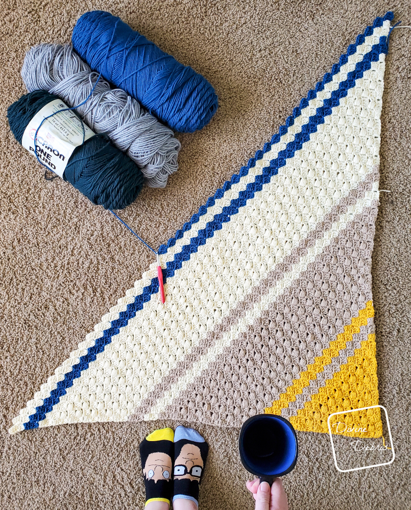 [Image description] A top down view of the unfinished Fitzgerald Blanket with 3 skeins of yarn along the top left of the blanket and a white woman's socked feet and a hand holding a cup of coffee in the bottom of the frame.