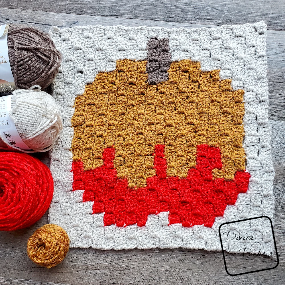 [Image description} finished C2C Caramel Apple Afghan Square laying flat on a wood grain background with skeins of yarn on the left side of the photo.