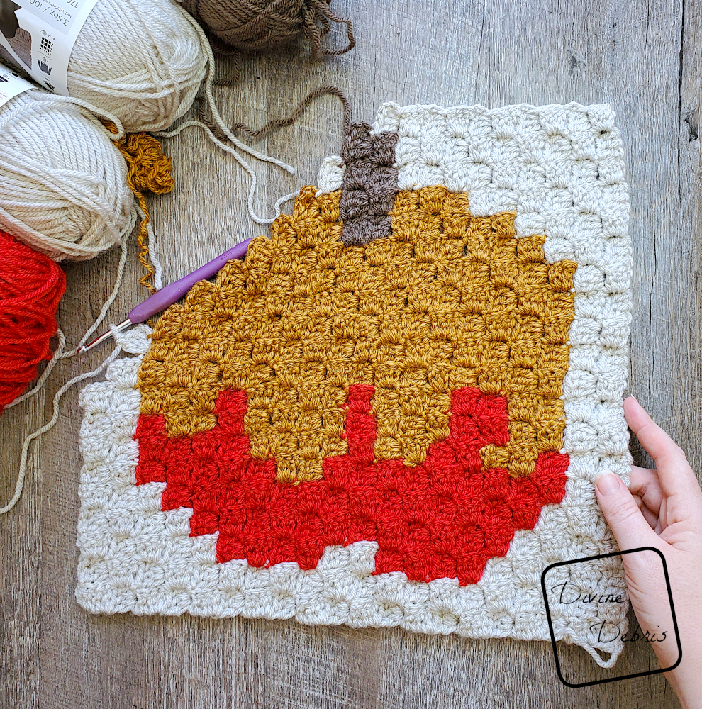 [Image description] A half-finished C2C Caramel Appe Afghan Square lays on a wood grain background, with a white woman's hand holding the right-hand side of the square. 