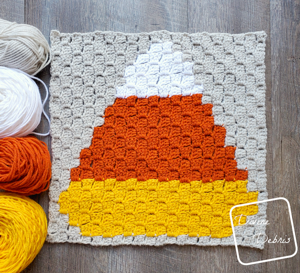 [Image description] C2C Candy Corn Afghan Square laying flat on a wood grain background with 4 skeins of yarn, along the left side of the square