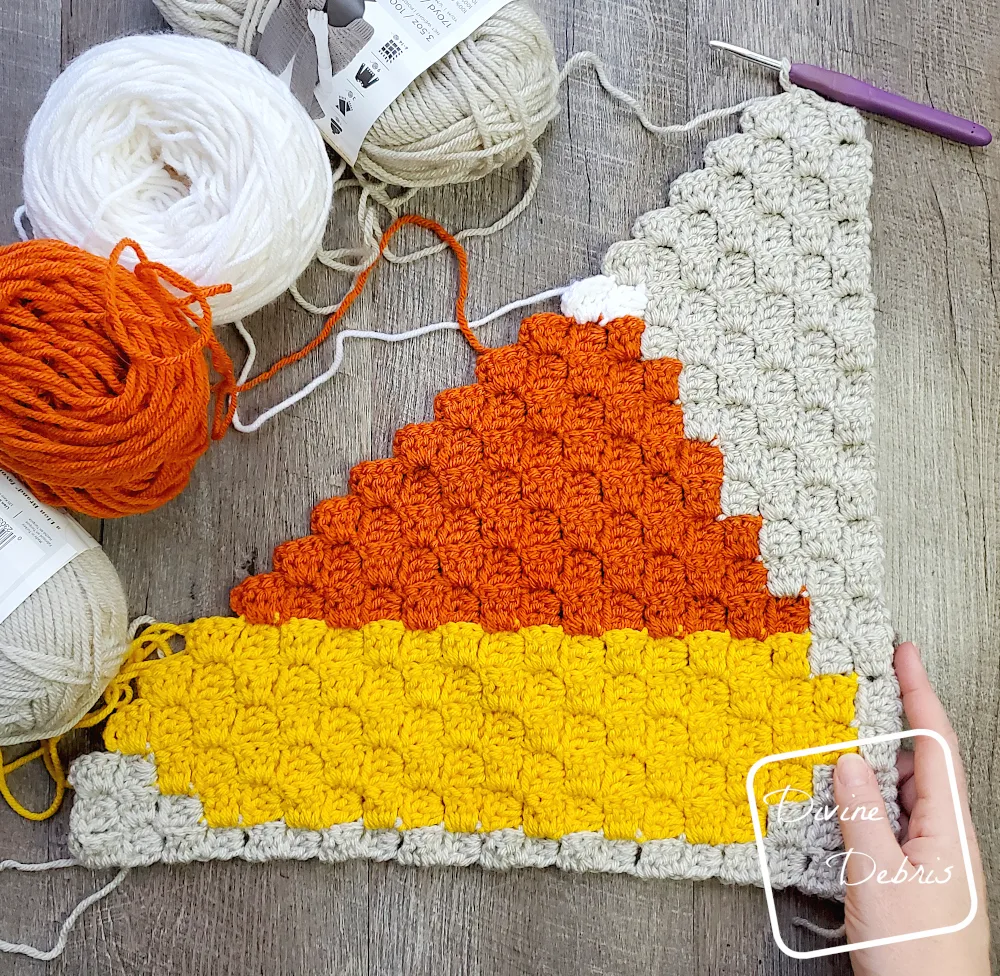 [Image description] A half-finished C2C Candy Corn Afghan Square lays flat on a wood-grain background, with skeins of yarn along the top left and a white woman's hand holding the bottom right corner.