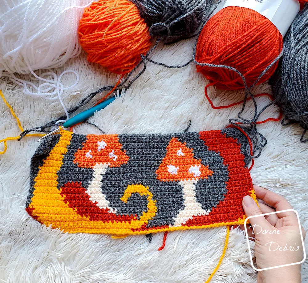 [Image description] Top down view of a quarter finished Ghost in the Mushrooms Wall Hanging on a white faux fur background. Skeins of yarn are visible at the top of the photo and a white woman's hand holds the bottom right corner.