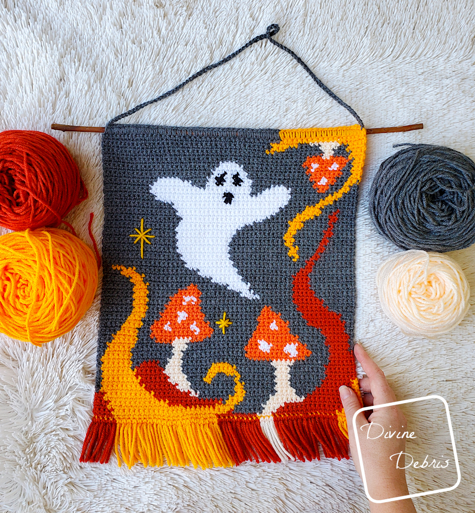 [image description] Top down view of the Ghost in the Mushrooms Wall Hanging laying on a white fur type blanket with 4 cakes of yarn on the sides and a white woman's hand holding the bottom right corner.