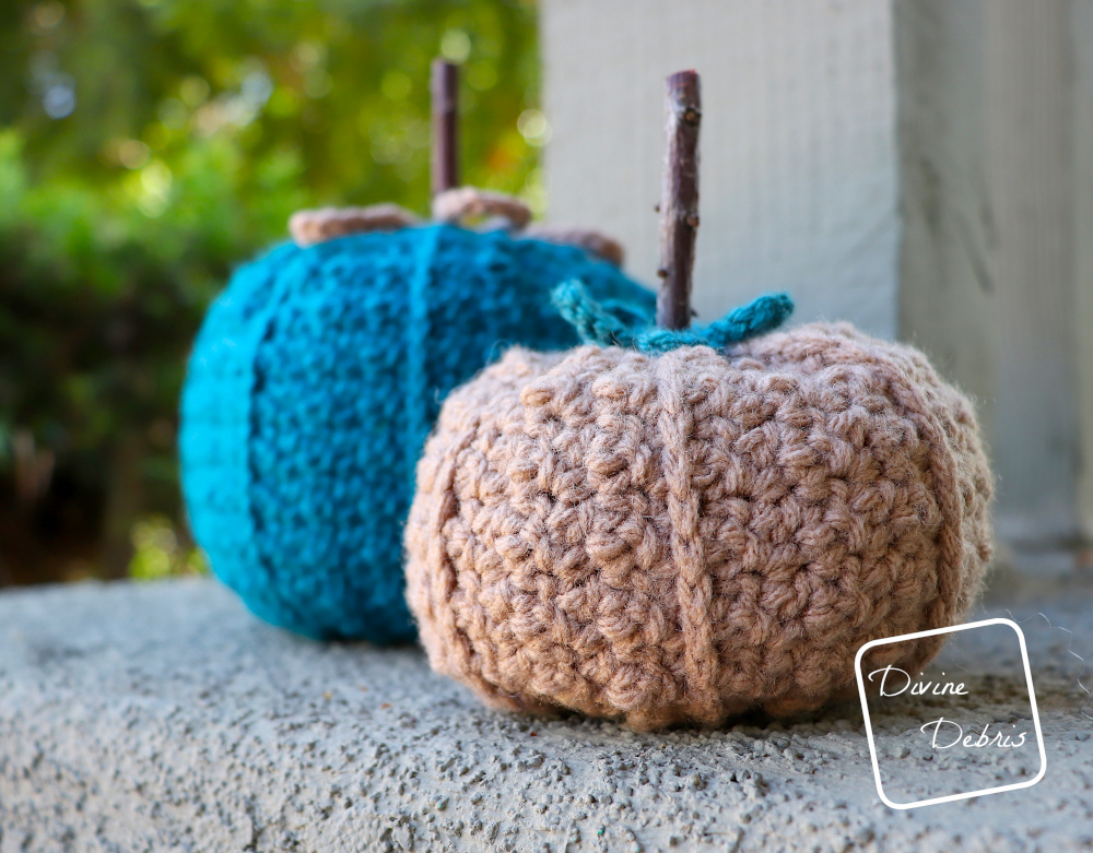 [Image description] the two sizes of the Kieran Pumpkin, the large one made in blue yarn sits in back and the small one made in brown yarn sits in front, on a platform in front of green bushes and sunlight
