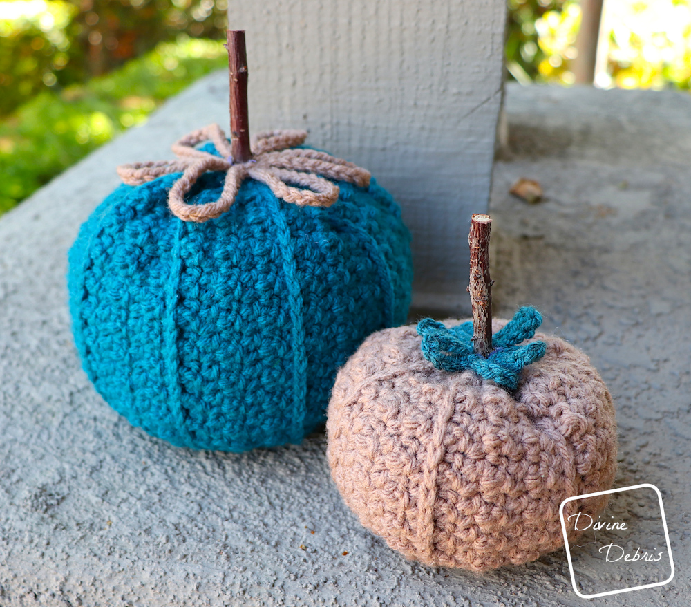 [Image description] the two sizes of the Kieran Pumpkin, the large one made in blue yarn sits in back and the small one made in brown yarn sits in front, on a platform in front of green bushes and sunlight
