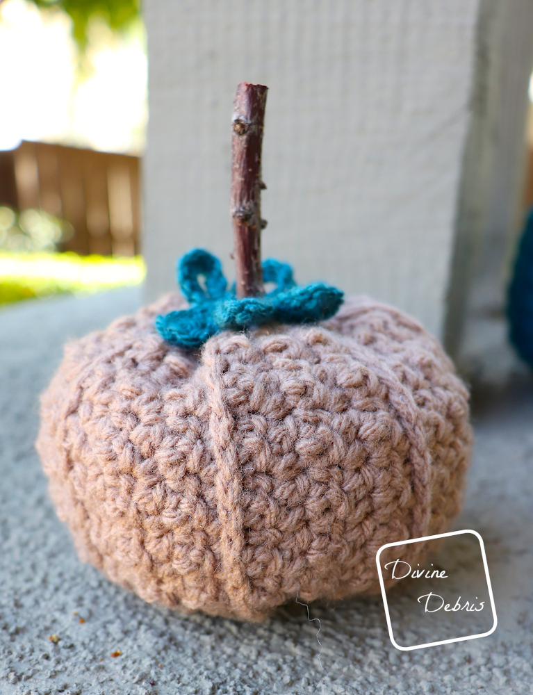 [Image description] the small Kieran Pumpkin made in brown yarn sits  on a platform in front of green bushes and sunlight