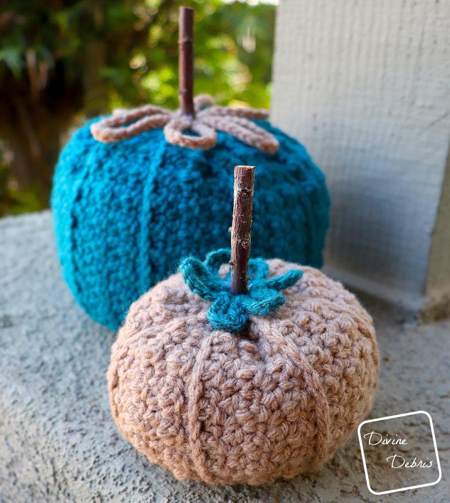[Image description] the two sizes of the Kieran Pumpkin, the large one made in blue yarn sits in back and the small one made in brown yarn sits in front, on a platform in front of green bushes 