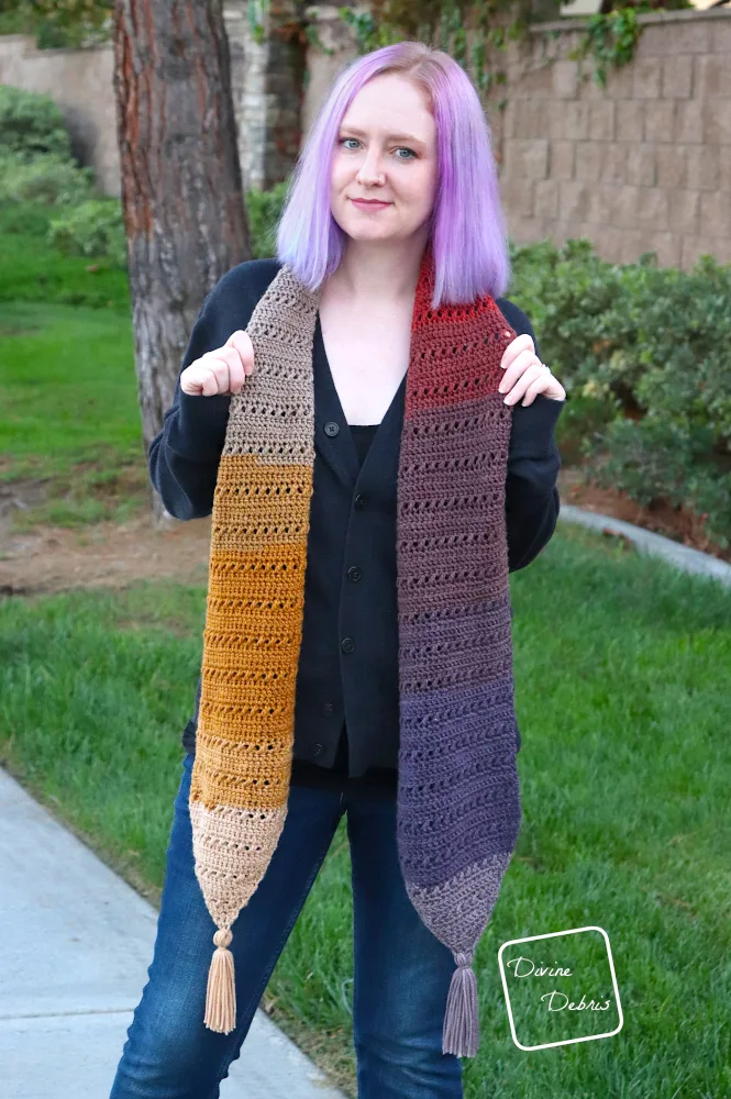 [image description] A white woman with purple hair stands in front of a tan wall and grass wearing the Sophie Scarf long way